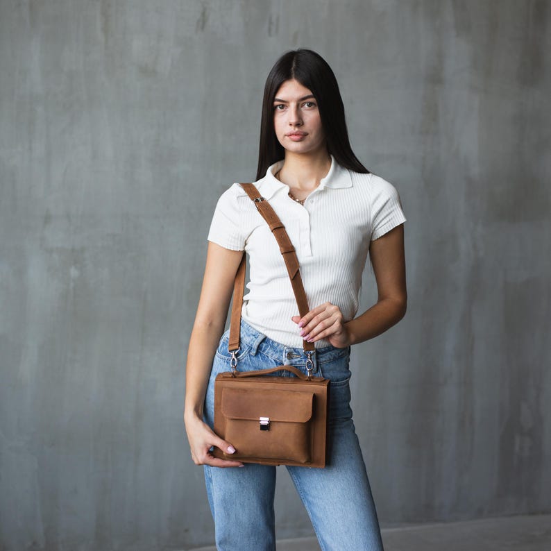 May include: A woman wearing a white collared shirt and blue jeans is carrying a brown leather satchel bag with a shoulder strap.