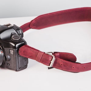 May include: A burgundy leather camera strap with a silver buckle. The strap is attached to a black Canon EOS 5D camera with a lens.