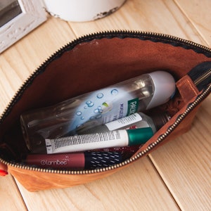 May include: A brown leather zippered pouch with a black zipper pull. The pouch is open and contains various toiletries, including a clear bottle of body wash, a tube of lip balm, and a small bottle of hand sanitizer.