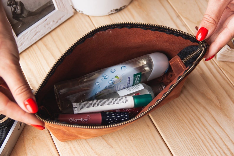 May include: A brown leather zippered pouch with a black zipper pull. The pouch is open and contains various toiletries, including a clear bottle of body wash, a tube of lip balm, and a small bottle of hand sanitizer.