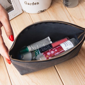 May include: A black leather zippered pouch with a gold zipper, open to reveal various beauty products, including a green tube, a white tube, a pink tube, and a clear bottle with a white label.