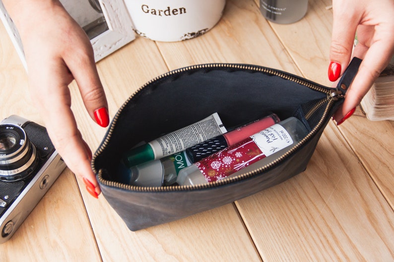 May include: A black leather zippered pouch with a gold zipper, open to reveal various beauty products, including a green tube, a white tube, a pink tube, and a clear bottle with a white label.