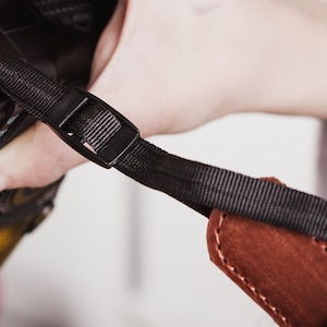May include: A black camera strap with a brown leather pad and a black plastic buckle.