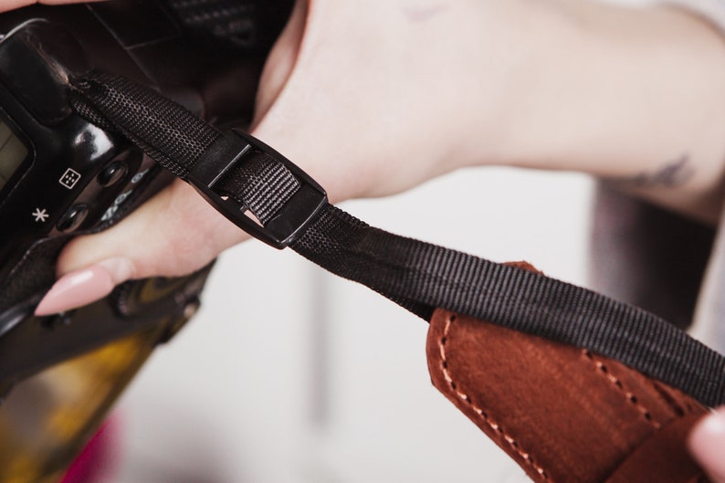May include: A black camera strap with a brown leather pad and a black plastic buckle.