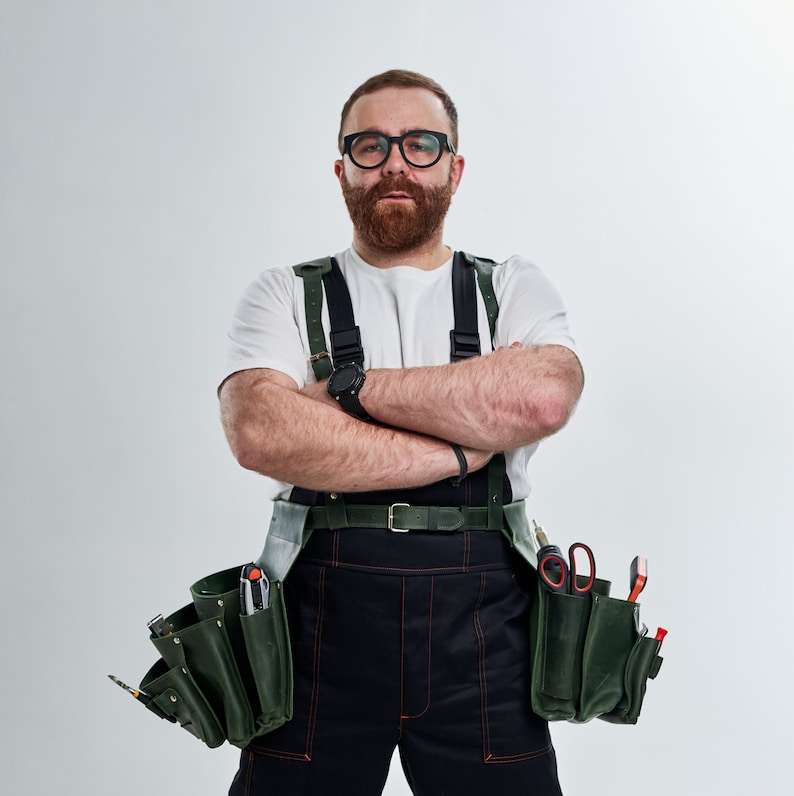 K&ouml;nnte beinhalten: Ein Mann tr&auml;gt ein wei&szlig;es T-Shirt, eine schwarze Arbeitshose und einen gr&uuml;nen Werkzeugg&uuml;rtel mit verschiedenen Werkzeugen. Er tr&auml;gt eine Brille und hat einen Bart. Er hat die Arme vor der Brust verschr&auml;nkt.