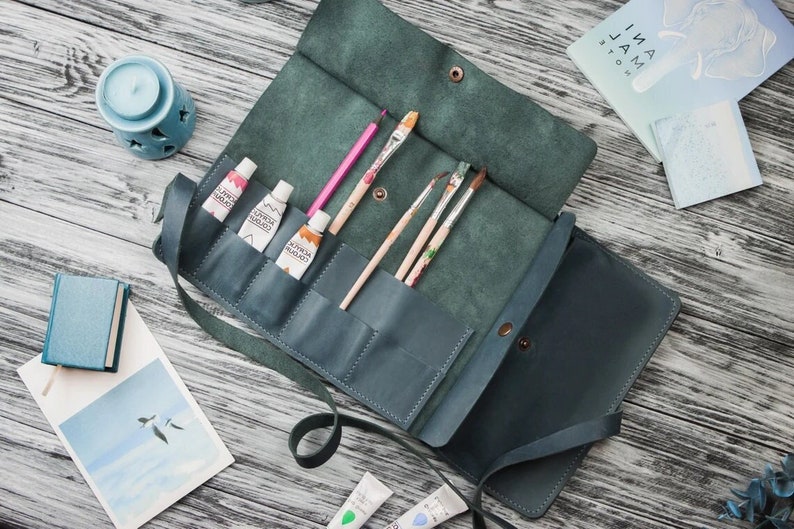 May include: A teal leather roll-up case with multiple pockets for paint tubes and brushes. The case has a strap and a flap closure. The case is open and the pockets are filled with paint tubes and brushes.