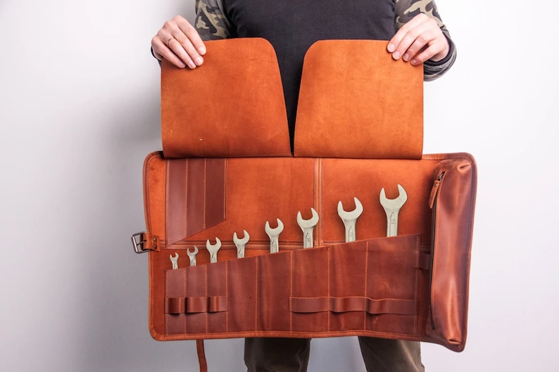 Peut inclure: Un rouleau &agrave; outils en cuir marron avec une poche zipp&eacute;e et plusieurs fentes pour ranger des cl&eacute;s. Le rouleau est ouvert et les cl&eacute;s sont visibles.