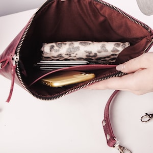 Peut inclure: Une pochette en cuir bordeaux avec une fermeture &eacute;clair et une bandouli&egrave;re amovible. La pochette est ouverte et montre un t&eacute;l&eacute;phone, un portefeuille et des cartes &agrave; l'int&eacute;rieur.