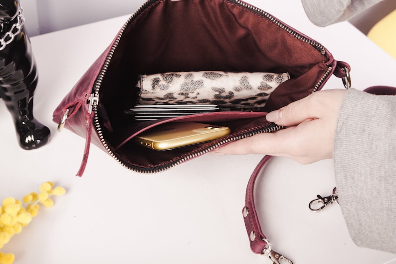 Peut inclure: Une pochette en cuir bordeaux avec une fermeture &eacute;clair et une bandouli&egrave;re amovible. La pochette est ouverte et montre un t&eacute;l&eacute;phone, un portefeuille et des cartes &agrave; l'int&eacute;rieur.