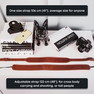 May include: Two brown leather camera straps, one 106 cm long and the other 122 cm long. The straps are laid out on a white surface with various designer books and a black ceramic dog figurine.