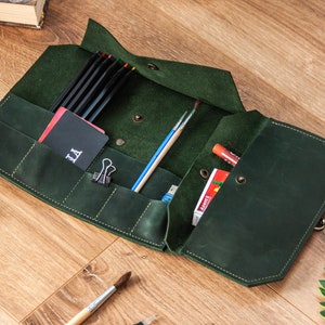 May include: A dark green leather roll-up case with multiple pockets and compartments. The case is open and contains pencils, brushes, and other art supplies. The case has a metal clasp and a small pocket with the word "LA" embroidered on it.