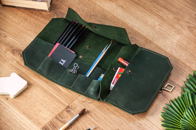 May include: A dark green leather roll-up case with multiple pockets and compartments. The case is open and contains pencils, brushes, and other art supplies. The case has a metal clasp and a small pocket with the word "LA" embroidered on it.