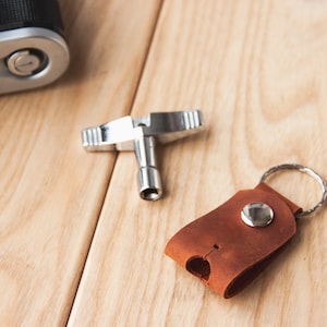 May include: A silver-toned camera lens wrench and a brown leather key chain with a silver ring and button.