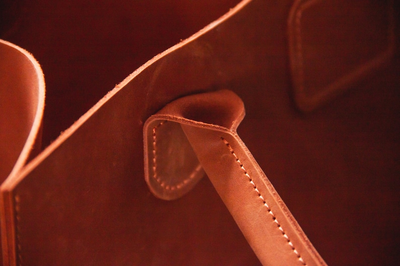 May include: Close-up of a brown leather tote bag with a stitched handle. The handle is folded over and stitched in place.