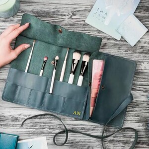 May include: Teal leather makeup brush roll with a zipper closure and multiple slots for brushes. The roll is open and has brushes inside. The roll is monogrammed with the initials "AN".