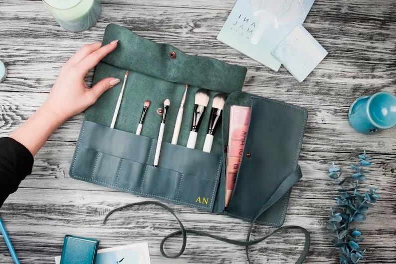 May include: Teal leather makeup brush roll with a zipper closure and multiple slots for brushes. The roll is open and has brushes inside. The roll is monogrammed with the initials "AN".