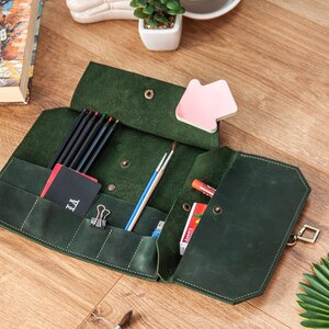 May include: A green leather pencil case with multiple pockets and compartments. The case is open and filled with coloured pencils, paintbrushes, and other art supplies. The case has a metal clasp.
