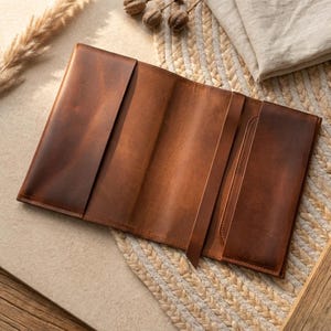 May include: Open, brown leather journal with a textured surface. The journal has multiple interior pockets and a leather strap closure. The journal is open, revealing the interior pages. The leather has a distressed appearance.