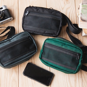 Könnte beinhalten: Drei kleine schwarze und grüne Canvas-Taschen mit verstellbaren Trägern. Die Taschen haben ein Reißverschlussfach und ein Netzfach. Die Taschen befinden sich auf einer Holzoberfläche.
