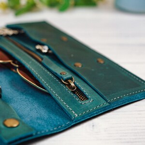 May include: A teal leather wallet with a zipper closure, a ring holder, and a glasses compartment. The wallet is open and the zipper is pulled down.