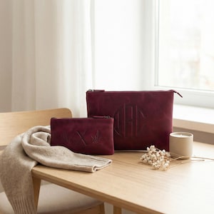 Pu&ograve; includere: Due pochette in pelle bordeaux con chiusure a zip. La pochette pi&ugrave; grande presenta un monogramma, mentre quella pi&ugrave; piccola mostra la lettera "X" con accenti di foglie. Una sciarpa beige e una candela sono anche sul tavolo di legno.
