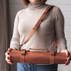 May include: A brown leather roll with a strap and buckles. The roll has a handle and is being held by a person.