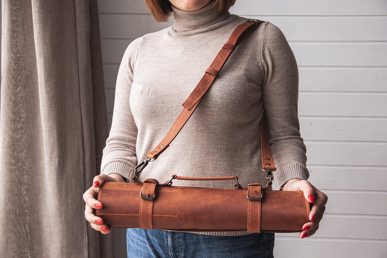 May include: A brown leather roll with a strap and buckles. The roll has a handle and is being held by a person.