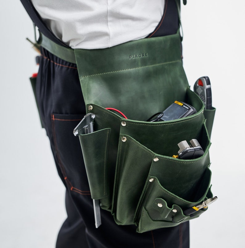 May include: A green leather tool belt with multiple pockets and compartments, worn over black work pants. The belt is labeled "PIKORE" and is filled with various tools, including a multimeter, wire cutters, and a screwdriver.