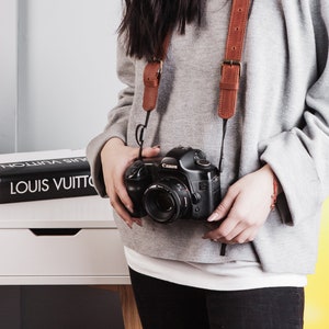 May include: A person wearing a grey jumper and black trousers is holding a black Canon camera with a brown leather camera strap. The camera is hanging around their neck.