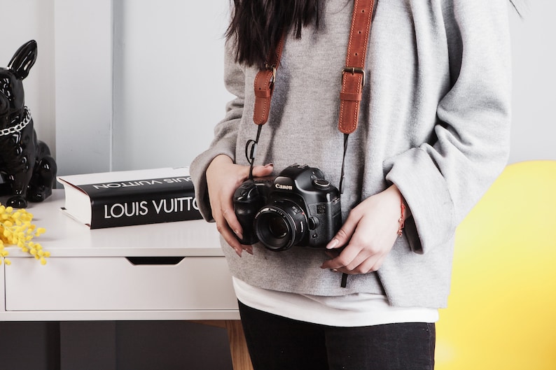 May include: A person wearing a grey jumper and black trousers is holding a black Canon camera with a brown leather camera strap. The camera is hanging around their neck.