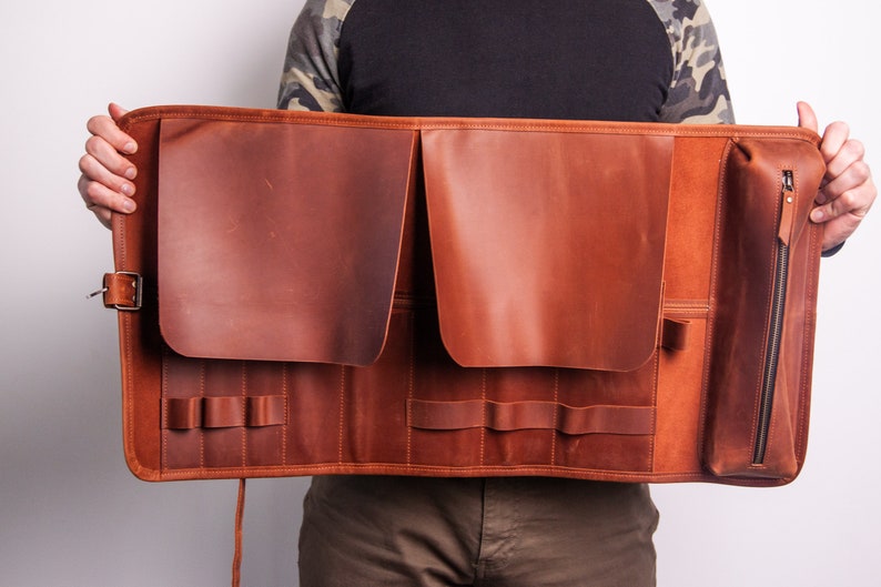 Peut inclure: Un &eacute;tui enroulable en cuir marron avec plusieurs poches et fentes pour ranger des outils ou d'autres objets. L'&eacute;tui a une poche zipp&eacute;e d'un c&ocirc;t&eacute; et une sangle avec une boucle de l'autre.