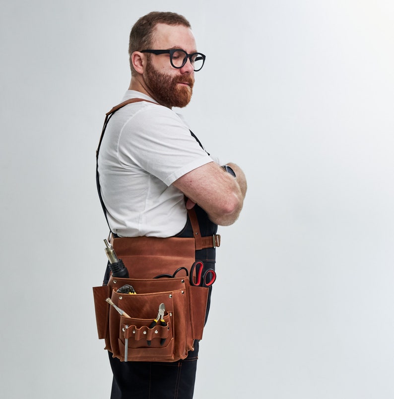 May include: A man wearing a white t-shirt and a brown leather tool belt with various tools and a pair of scissors. He is standing with his arms crossed and looking down.