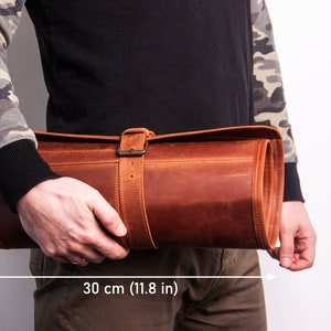 Peut inclure: Un sac enroulable en cuir marron avec une fermeture &agrave; boucle. Le sac mesure 30 cm de large et 13 cm de haut.