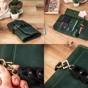 May include: A green leather pencil case with a black strap and brass hardware. The case is open and shows multiple compartments for pencils, pens, and other writing tools.