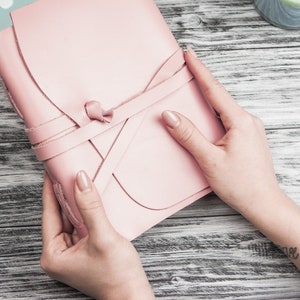 May include: A pink leather journal with a tied closure. The journal is being held by two hands.