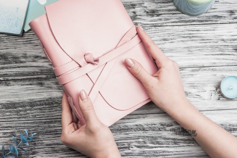 May include: A pink leather journal with a tied closure. The journal is being held by two hands.