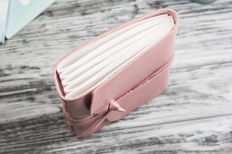 May include: A pink leather-bound journal with blank pages. The journal is closed and lying on a wooden surface.