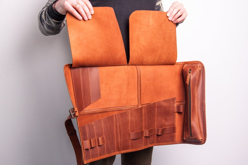 Peut inclure: Un &eacute;tui enroulable en cuir marron avec plusieurs compartiments et une poche zipp&eacute;e. L'&eacute;tui est ouvert pour r&eacute;v&eacute;ler l'int&eacute;rieur, qui comporte plusieurs fentes pour ranger des outils ou d'autres objets.