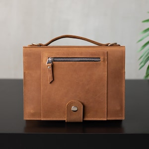 May include: A brown leather briefcase with a zippered pocket and a strap closure. The briefcase has a top handle and a flap closure.