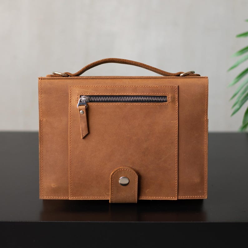 May include: A brown leather briefcase with a zippered pocket and a strap closure. The briefcase has a top handle and a flap closure.