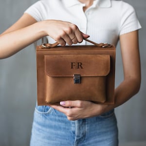 以下が含まれることがあります： 前面に「F|R」の刻印が施された、銀色の留め具が付いた茶色の革製ブリーフケースです。