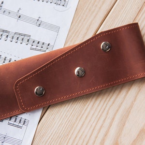 May include: A brown leather strap with three silver snaps. The strap is lying on a wooden surface with sheet music underneath.