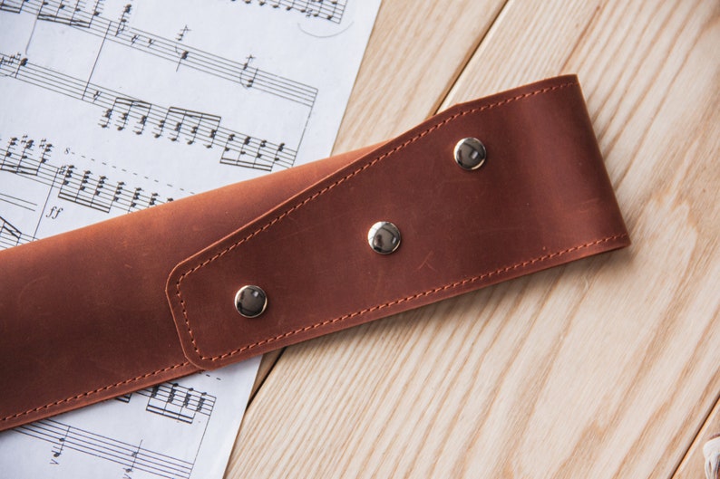 May include: A brown leather strap with three silver snaps. The strap is lying on a wooden surface with sheet music underneath.