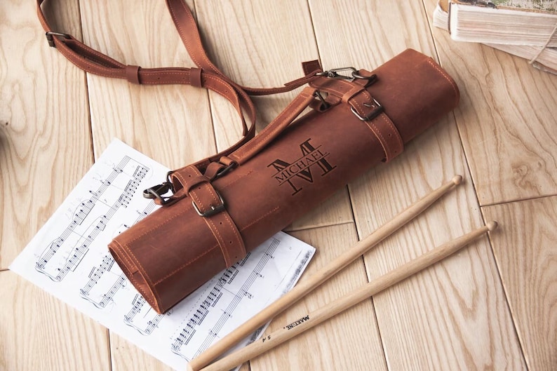 May include: A brown leather drumstick roll with a strap and buckle closure. The roll is personalised with the name "Michael" and the initial "M". Two wooden drumsticks are lying next to the roll on a wooden surface.