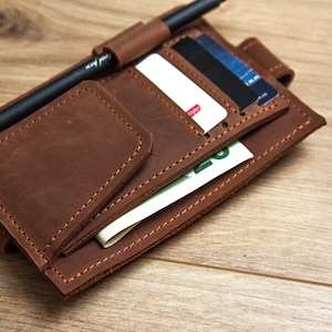 May include: A brown leather wallet with multiple pockets and slots for cards, cash, and a pen. The wallet is closed and lying on a wooden surface.