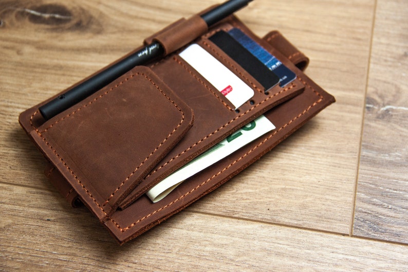 May include: A brown leather wallet with multiple pockets and slots for cards, cash, and a pen. The wallet is closed and lying on a wooden surface.