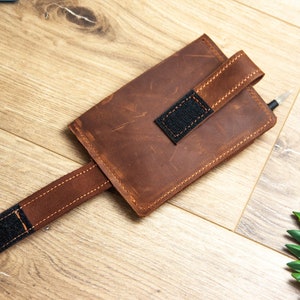 May include: A brown leather phone case with a strap and a black velcro closure. The case is on a wooden surface.