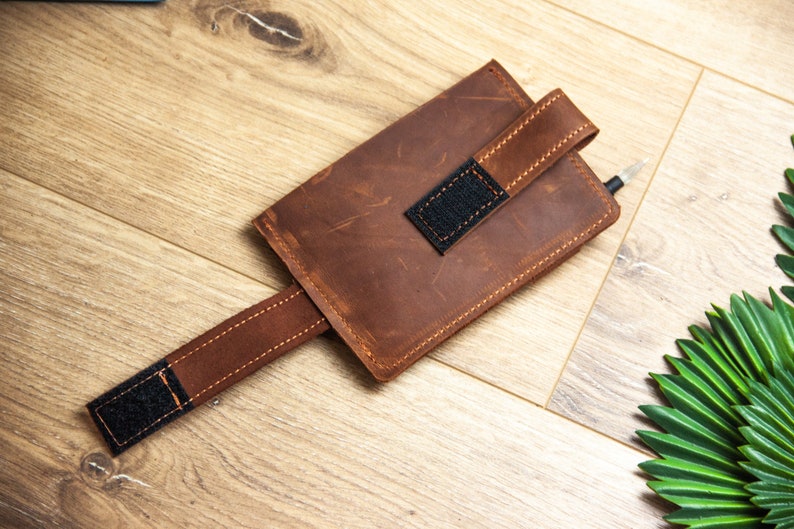 May include: A brown leather phone case with a strap and a black velcro closure. The case is on a wooden surface.