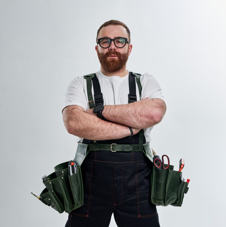May include: A man wearing a white t-shirt, black work pants, and a green tool belt with various tools. He is wearing glasses and has a beard. He has his arms crossed in front of him.