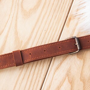 May include: A brown leather belt with a silver buckle. The belt is stitched with brown thread and has multiple holes for adjusting the size.